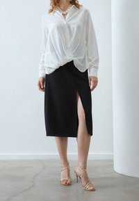 Kvinna i vit långärmad blus, svart midiklänning med sidöppning, nude strappy högklackade sandaler, står på grått golv vid vit vägg.