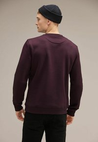 Felpa bordeaux con scollatura rotonda, polsini e orlo a coste, e design semplice. Indossata con pantaloni neri e un berretto scuro.