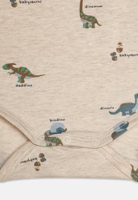 Bambusova bodysuit v svetlo bežovi z barvnimi potiski dinozavrov. Ima pritisne zasnove na dnu in igrive tekstovne oznake ob vsakem motivu.