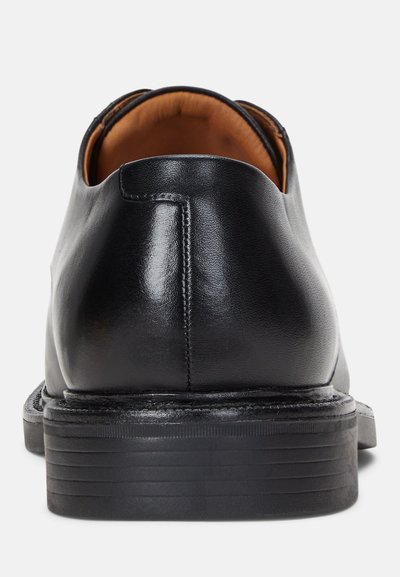 Zapato de cuero negro con parte trasera redondeada, textura suave y detalles de costura sutiles. La suela está ligeramente elevadas con acentos estriados.