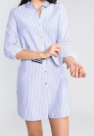 Kobieta poprawiająca rękaw sukienki koszulowej w pionowe niebiesko-białe paski z czarno-białym paskiem w talii.