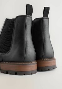 Fekete bőrből készült bokacsizma pár, szőtt oldalsó panelek, barna réteges sarkak és húzófülek jellemzik, világos háttér előtt.