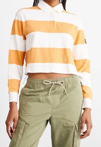 Gestreiftes Langarm-Poloshirt in Orange und Weiß mit Kragen und Knopfleiste, kombiniert mit olivgrünen Cargo-Hosen mit Kordelzug.