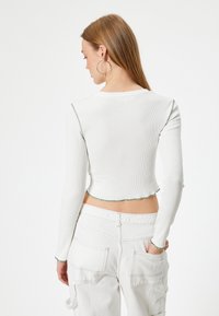 Witte langmouwige cropped top met ribbelstructuur en groene contraststiksels langs de naden en de zoom, gecombineerd met witte cargobroek.
