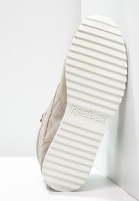 Sneaker with a soft beige suede upper, white rubber sole featuring horizontal stripes, and embossed Reebok logo. Side zipper detail visible.