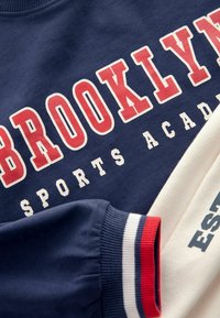 Granatowy sweter z nadrukiem "BROOKLYN SPORTS ACADEMY" w intensywnych barwach czerwonej i białej. Żebrowane mankiety z paskami w kolorach czerwonym, białym i niebieskim. Miękka faktura.
