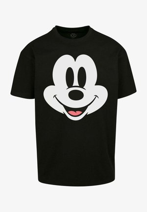 T-shirt en coton noir avec un grand imprimé de visage de dessin animé blanc. Associé à un pantalon en denim foncé et des baskets blanches, se tenant devant un fond clair.