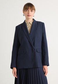 Námořnický blazer s pruhy a klopovými límci, se dvěma předními kapsami a volným střihem, doplněný světlým béžovým košilí s límcem.