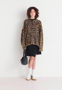 Pull oversized à imprimé léopard en beige et marron, jupe mini noire, chaussettes blanches et baskets. Un sac noir finit la tenue.