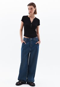 Sort kortærmet henley-top med tre knapper; brede blå denimjeans; sorte sandaler med en tyk sål. Modellen har kort hår.