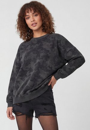 Ung kvinde med krøllet hår iført en mørkegrå camouflage-sweatshirt, sorte ødelagte shorts og sorte strømpebukser, poserer med den ene hånd på hoften.