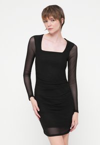 Rochie neagră cu mâneci lungi din țesătură transparentă, decolteu pătrat și textură încrețită. Designul aderent subliniază silueta.