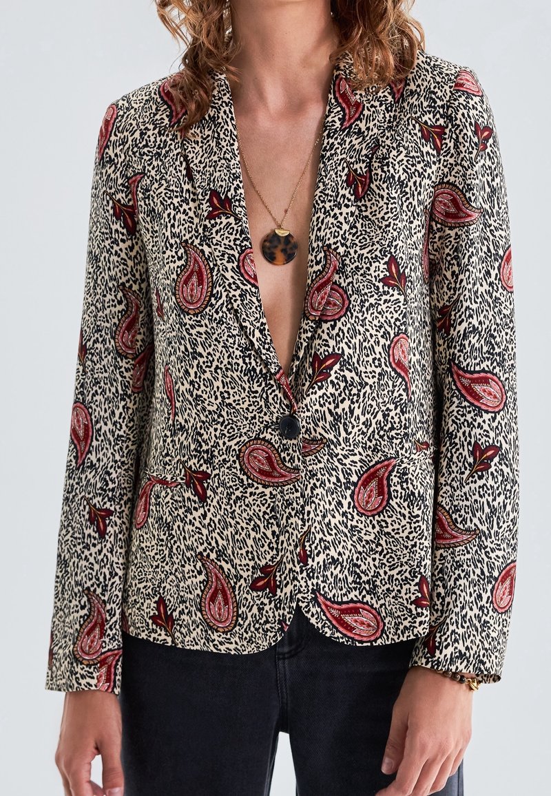 Tryckt kavaj i krämfärg med röd paisley- och svartprickigt mönster. Har ett snitt med slag, knappstängning och långa ärmar.