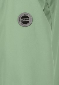 Mintgrön tyg med en slät yta som har ett runt svart och silverfärgat logomärke med texten "ZIGZAG" på vänster sida.