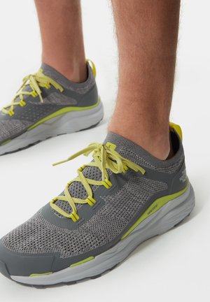 Zapatillas deportivas grises con parte superior de malla, acentos amarillos y cordones. La suela es blanca con un patrón de agarre texturizado y un talón prominente.