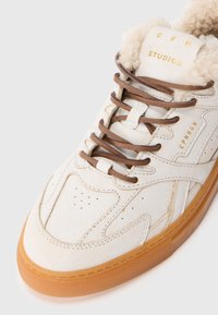 Zapatillas de cuero crema con cordones marrones, suela de goma, forro de borreguito y marca dorada "CPH STUDIOS" en la lengüeta y el lateral.