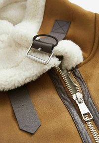 Gros plan d'une veste en shearling marron avec fermeture éclair argentée, sangle de boucle en cuir marron foncé et doublure blanche duveteuse.
