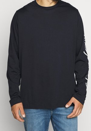 Uomo che indossa una maglia nera a maniche lunghe con scritte bianche sulla manica destra e jeans blu, in piedi davanti a uno sfondo semplice.