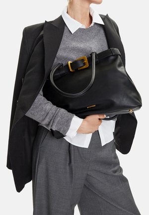 Person in grauem Pullover, weißem Hemd, schwarzem Blazer und grauer Hose, die eine große schwarze Ledertasche mit goldener Schnalle hält.