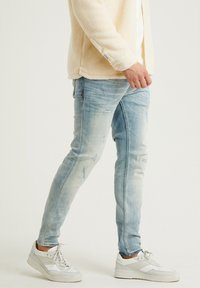 Vaqueros ajustados de azul claro con detalles desgastados, con bolsillos delanteros y un lavado desgastado, combinados con zapatillas blancas.