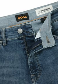 Jeans de denim en un lavado oscuro con una textura desgastada. Presenta un cierre de cremallera, botón y etiquetas de marca BOSS.