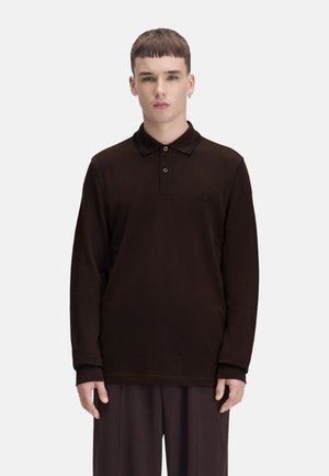 Joven de pie de frente vistiendo una camisa polo negra de manga larga y pantalones oscuros a juego sobre un fondo liso.