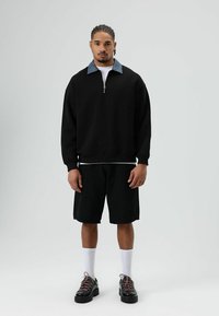 Zwarte fleece trui met een denim kraag, gecombineerd met zwarte korte broek. Draagt witte sokken en zwarte schoenen met decoratieve veters.