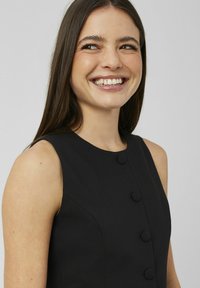 Robe noire sans manches avec un col haut, comportant cinq boutons recouverts de tissu à l'avant et une coupe ajustée. Texture lisse.