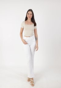 Femme aux longs cheveux noirs portant un top beige à encolure carrée, un pantalon blanc taille haute, des sandales et un tatouage floral sur l'avant-bras gauche.