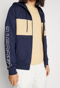 Felpa con zip di colore blu navy e beige, caratterizzata da un design bicolore, tasche frontali, cappuccio con coulisse e grande logo sulla manica. Texture liscia.