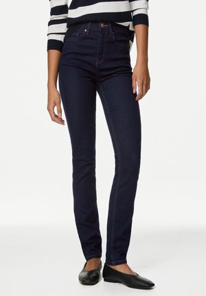 Person iført mørkeblå slim-fit jeans, en hvid og marineblå stribet sweater samt sorte flade sko, stående mod en ensfarvet baggrund.