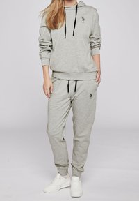 Kvinna klädd i en ljusgrå hoodie och matchande sweatpants med svarta dragsnören och vita sneakers, stående med ena handen i fickan.