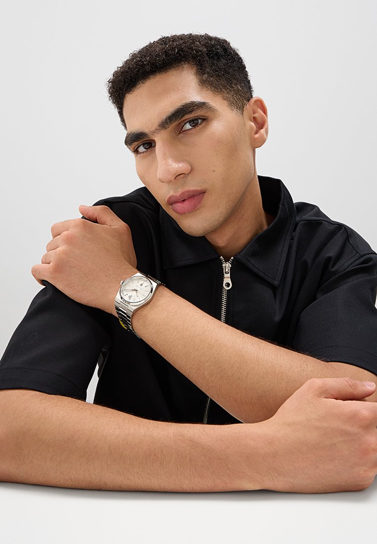 Montre argentée avec un cadran rond, bracelet noir avec un motif rayé, portée au poignet. Le modèle porte une chemise zippée noire.