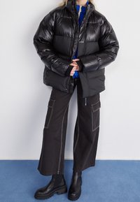 Black puffer jacket with a shiny finish, featuring a high collar and elastic cuffs. Paired with loose black pants and chunky black boots.