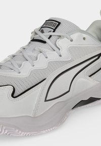 Λευκό και γκρι αθλητικό παπούτσι Puma με πλέγμα ύφασμα, μαύρες λεπτομέρειες με το λογότυπο και λευκά κορδόνια πάνω σε υφή σόλας.
