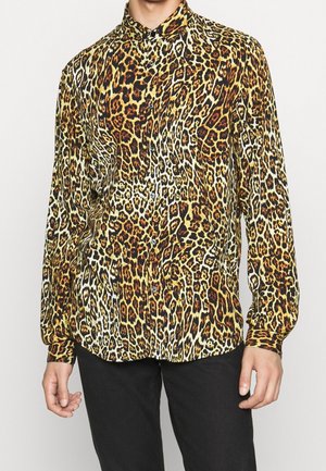 Homme portant une chemise à manches longues boutonnée avec un motif léopard jaune, marron et noir et un pantalon noir, debout devant un fond uni.