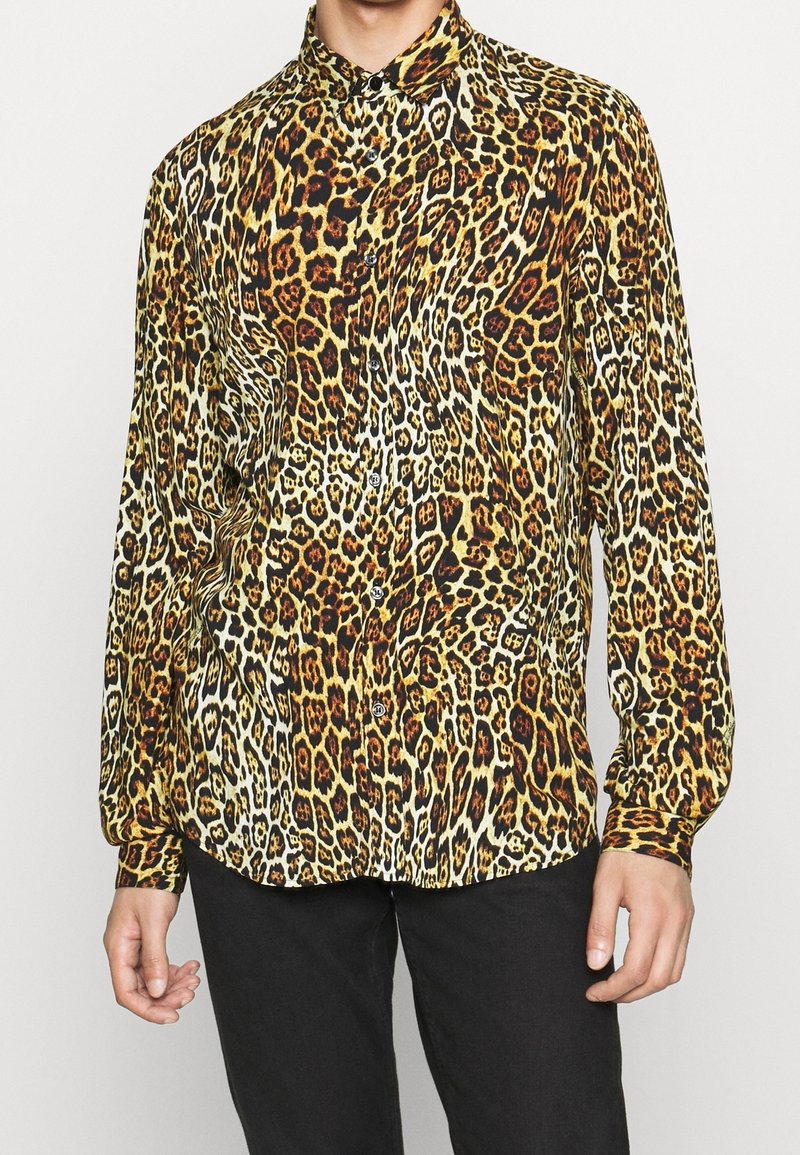 Homme portant une chemise à manches longues boutonnée avec un motif léopard jaune, marron et noir et un pantalon noir, debout devant un fond uni.