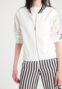 Bílé krajkové bomber bundě se zapínačkou, s žíhaným černobílým límcem a ohrnutými rukávy. V kombinaci s vertikálně pruhovanými černobílými kalhotami.