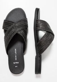 Svart flätad slide-sandal med korsade remmar, platt sula, texturerad yta och tryckt logotyp på innersulan.