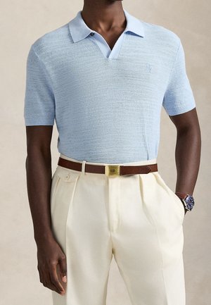 Homme portant un polo en maille bleu clair à manches courtes, rentré dans un pantalon plissé crème avec ceinture marron et montre-bracelet.