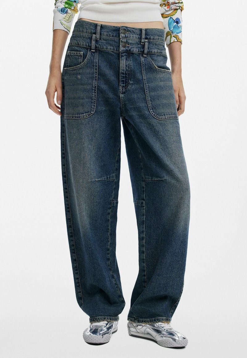 Jeans en denim foncé à jambes larges avec grandes poches frontales, portés par une personne chaussée de baskets argentées brillantes et portant un haut à manches blanches à motifs floraux.