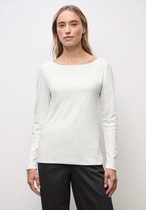 Mujer de pie contra un fondo liso, vestida con una camiseta blanca ceñida de manga larga y pantalones gris oscuro, mirando hacia adelante con una postura relajada.