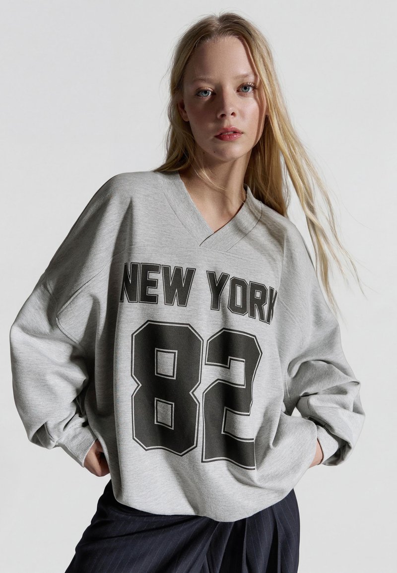 Młoda kobieta o długich blond włosach, ubrana w luźną szarą bluzę z napisem "NEW YORK 82" w czerni, pozująca z rękami w kieszeniach.