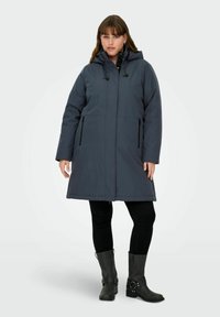 Sieviete, valkājot tumši pelēku kapuci ar priekšējām rāvējslēdzēm ziemas mēteļa, melnas legingus un melnas zābakus līdz vidus teļam, stāvot pret vienkāršu fonu.