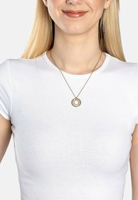 Guldtonet halskæde med en cirkulær vedhæng, der indeholder en central klar sten, båret over en tætsiddende hvid t-shirt. Simpelt design og elegante detaljer.