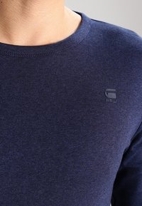 Detailní záběr na osobu v námořnické modré triku s kulatým výstřihem, na horní části hrudníku s malým logem G-RAW.