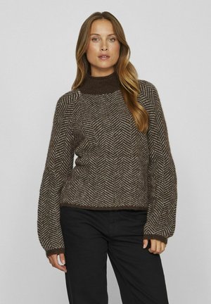 Brauner Pullover mit Fischgrätmuster, hohem Kragen und langen Ärmeln, mit strukturiertem Strick. Kombiniert mit schwarzen Hosen.