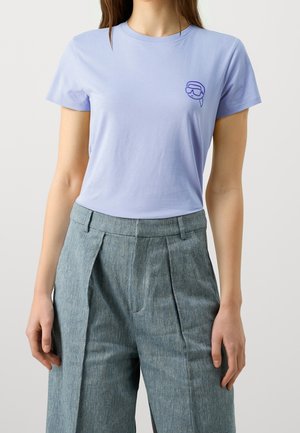 Žena ve světle fialovém tričku s krátkými rukávy, na kterém je malý obrázek šnorchlovací masky, a ve vysokým pasem šedých plisovaných kalhotách.