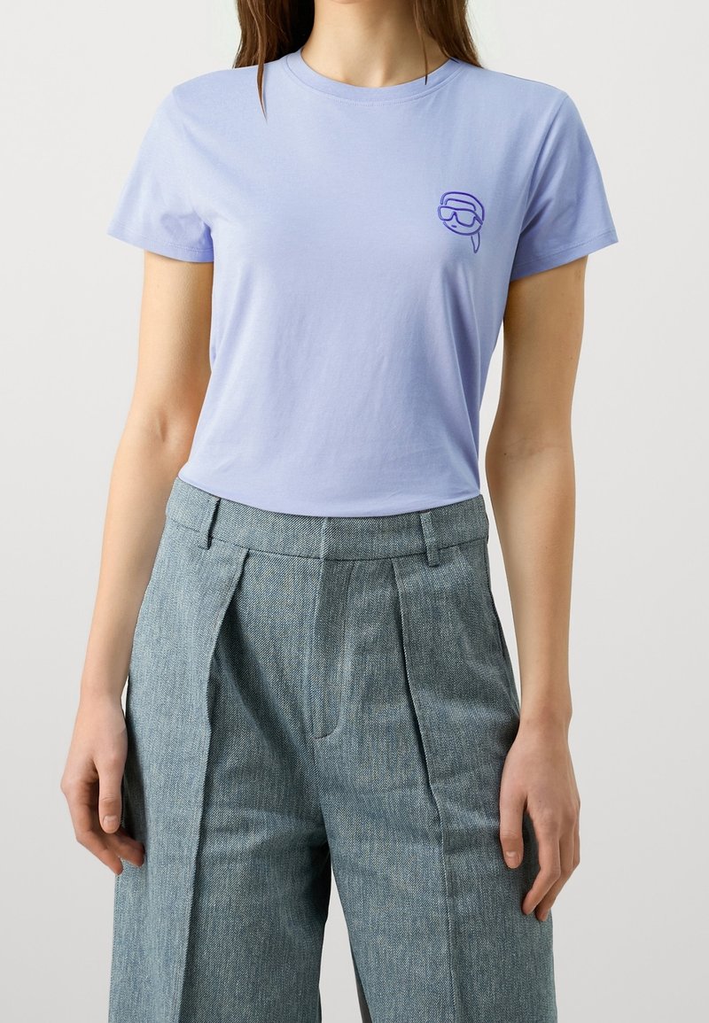 Kobieta nosząca jasnofioletową koszulkę z krótkim rękawem z małą grafiką maseczki do nurkowania i szare plisowane spodnie z wysokim stanem.