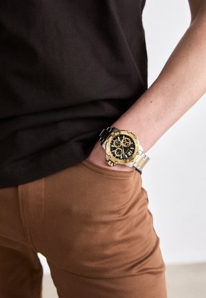 Reloj de pulsera dorado y negro con esfera redonda, características de cronógrafo y correa de metal. Pantalones marrones y camisa oscura visibles en primer plano.